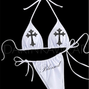 Puede incluir: Conjunto de bikini blanco con top halter y braguita con lazos. El top tiene dos diseños de cruz negra, y la braguita está impresa con la palabra "Blessed". El conjunto está hecho de una tela suave y ligera.