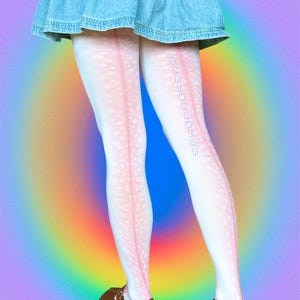 May include: White tights featuring a subtle pink speckled pattern and a vertical pink stripe running down the back of each leg. The text DARINKAWEAR is printed vertically in light blue letters on the left leg. A blue denim skirt and brown shoes.