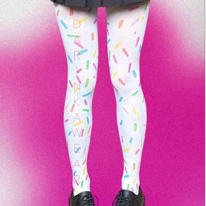 Puede incluir: Medias blancas con un patrón de confeti colorido y el texto "DARKWEAR" en letras verticales. La persona lleva zapatos negros y una falda plisada negra. El fondo es un degradado de rosa.