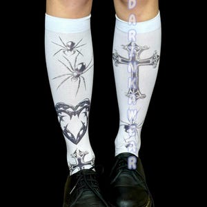 Puede incluir: Calcetines blancos hasta la rodilla con gráficos negros de arañas, corazones y cruces. La palabra "DARKWAKER" está impresa verticalmente en los calcetines. Los calcetines se usan con zapatos de cuero negro.