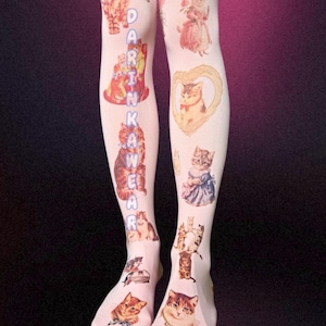 May include: White tights featuring a repeating pattern of various cat illustrations, including kittens in dresses and framed portraits. The text DARINKAWEAR is printed vertically in light blue letters along the side of the left leg.
