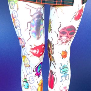May include: White leggings featuring a colorful all-over print of various beetles and insects in shades of red, blue, green, and yellow. The text DARINKAWEAR is printed vertically in light blue along the left leg. A green plaid skirt is visible at the top.