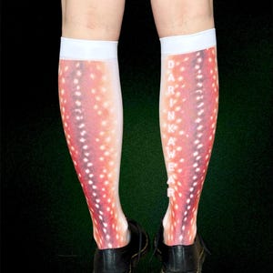 May include: Knee-high socks with a deer-like pattern in shades of red, pink, and white. The socks have a white band at the top and the word "DARKWEAR" printed vertically on the side. The socks are worn with black shoes.