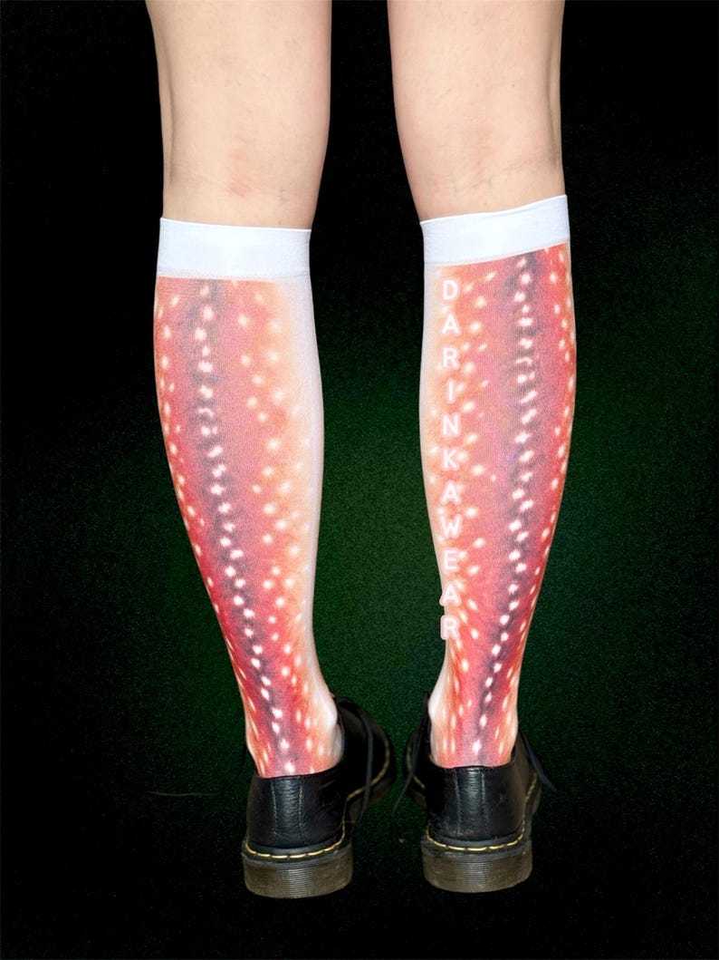 May include: Knee-high socks with a deer-like pattern in shades of red, pink, and white. The socks have a white band at the top and the word "DARKWEAR" printed vertically on the side. The socks are worn with black shoes.