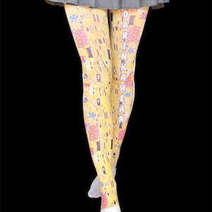 Peut inclure: Leggings jaunes avec un motif abstrait répétitif de formes dorées, roses et bleues, avec le texte vertical DARINKAWEAR imprimé en blanc sur la jambe droite, portés sous une jupe plissée grise sur un fond noir uni.
