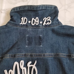 May include: A blue denim jacket with white embroidery. The back of the jacket says "Mrs." and "10.09.23".
