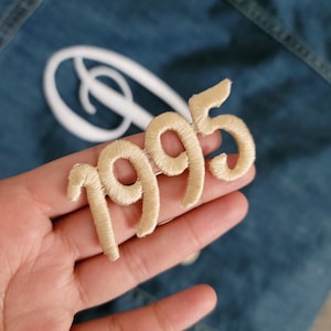 Könnte beinhalten: Ein beigefarbener gestickter Aufnäher mit dem Jahr 1995 in einer kursivschrift.