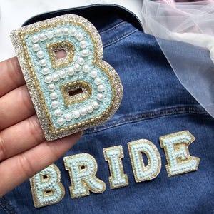 Puede incluir: Una chaqueta vaquera con fondo azul claro y una gran letra B azul claro brillante con detalles de perlas blancas. La palabra "BRIDE" está escrita en letras azul claro brillantes más pequeñas con detalles de perlas blancas.