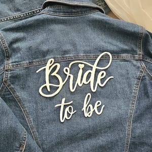 以下が含まれることがあります： 背面に「Bride to be」と白い筆記体で書かれた青いデニムジャケット。テキストの「i」の文字の上には小さなハートのアイコンがあります。ジャケットにはクラシックな襟、目に見える縫い目、フロントボタンが付いています。