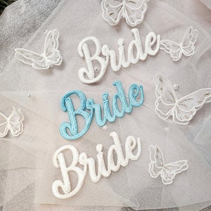 May include: Three white embroidered patches with the word "Bride" on a white tulle fabric background. The patches are in different sizes and fonts. There are also several white embroidered butterflies on the fabric.