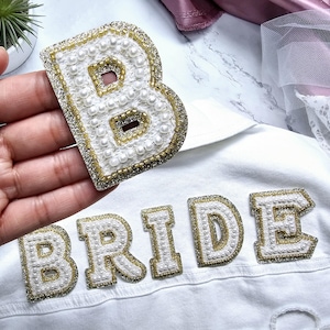 Peut inclure: Veste en jean blanche avec le mot "BRIDE" écrit en perles blanches et dorées. Une grande lettre "B" en perles blanches et garniture dorée se trouve sur le côté gauche de la veste.