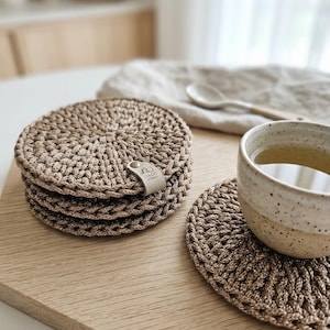 Può includere: Una pila di quattro sottobicchieri rotondi all'uncinetto color beige con un motivo intrecciato e testurizzato. Una piccola etichetta in pelle chiara è fissata sul lato della pila, con il testo in rilievo LU DECO STUDIO. Un sottobicchiere singolo è accanto.