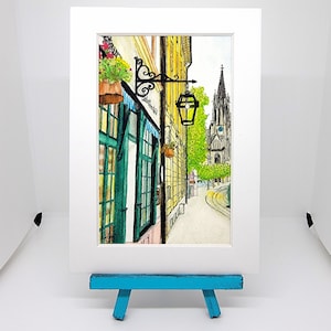 May include: Watercolor painting of a European street scene. The artwork features a building with green doors and a blue awning, a street lamp, and a church in the background. The painting is displayed on a white mat and a small blue easel.