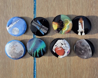May include: A collection of eight round space-themed buttons. The buttons feature illustrations of a rocket, a satellite, a space shuttle, planets, and an astronaut holding a red planet. The buttons are black with colorful designs.