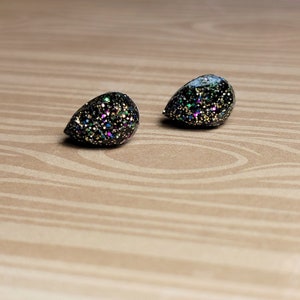 May include: Two teardrop-shaped earrings with a black base and colorful glitter embedded throughout. The earrings are displayed on a light brown wooden surface. The glitter includes gold, green, blue, and purple specks.