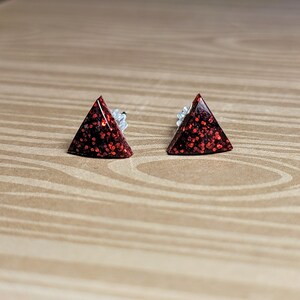 May include: A pair of red glitter triangle stud earrings. The earrings are small and have a simple design.