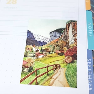 May include: A watercolor painting of a mountain village scene. The artwork depicts a path leading to a village with houses and a church, surrounded by green fields, trees, and mountains. The color palette includes greens, browns, and blues.