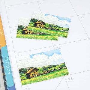May include: Three watercolor stickers featuring a green field with a brown house, green trees, and blue sky with white clouds. The stickers are placed on a white calendar with the numbers 10, 17, and 18.