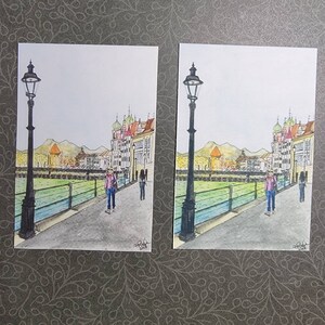 May include: A watercolor painting of a city scene with buildings, a bridge, and two people walking. The painting is done in shades of blue, green, and brown. There is a lamppost on the left side of the painting.
