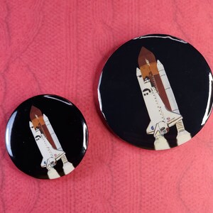 May include: Two round black buttons with a white space shuttle design. The larger button shows the space shuttle in flight with a black background. The smaller button shows the space shuttle in flight with a black background.