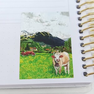 May include: A watercolor painting of a cow in a field with mountains in the background. The cow is brown and white with a bell around its neck. There are other cows in the distance and green grass and yellow flowers in the field.
