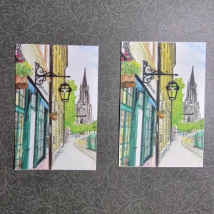 May include: A watercolor painting of a street scene with a tall church building in the background. The street is lined with buildings with green doors and windows. There are streetlights and a sign that says "Schnitzelwirt".