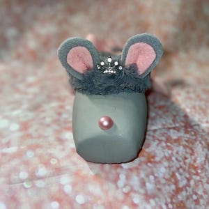 May include: A gray mouse figurine with large gray ears lined with pink, a fluffy gray headpiece, and a small jeweled crown. The mouse has a pink pearl nose. The figurine is set against a pink and white glittery background.