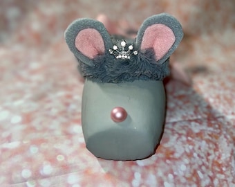 May include: A gray mouse figurine with large gray ears lined with pink, a fluffy gray headpiece, and a small jeweled crown. The mouse has a pink pearl nose. The figurine is set against a pink and white glittery background.