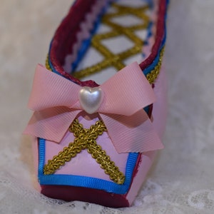 May include: A pink ballet shoe with a gold trim and a pink bow with a white heart-shaped embellishment.