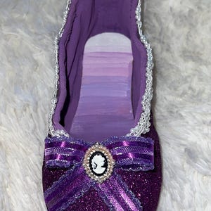 May include: A decorative, glittery purple ballet slipper with a bow and cameo. The slipper is embellished with a white beaded trim around the opening. The bow features a cameo with a white silhouette.
