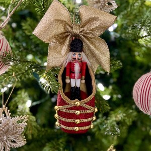 May include: A Christmas tree ornament featuring a red and gold nutcracker soldier inside a red boot-shaped decoration. The ornament is adorned with gold glitter and a large gold bow. Other Christmas ornaments are visible in the background.