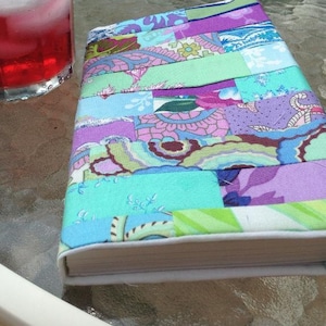 Puede incluir: Un libro hecho a mano con una cubierta de patchwork colorida hecha de diferentes telas en tonos de morado, azul, verde y rosa. La cubierta está hecha de tiras de tela cosidas juntas para crear un diseño único.