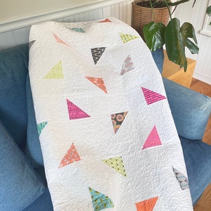 May include: A white quilt with a patchwork design featuring colorful triangles. The quilt is made with a variety of fabrics, including floral prints, geometric patterns, and solid colors. The quilt is displayed on a blue couch.