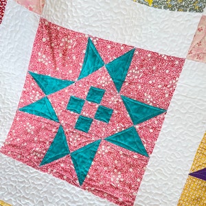 May include: A close-up of a quilt block with a teal star design on a pink background. The star is made up of eight triangles and is surrounded by a white border.