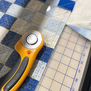 May include: An orange and black rotary cutter with a clear ruler is positioned on a blue and white checkered quilt. The ruler has measurements in centimetres. The quilt is in the process of being cut.