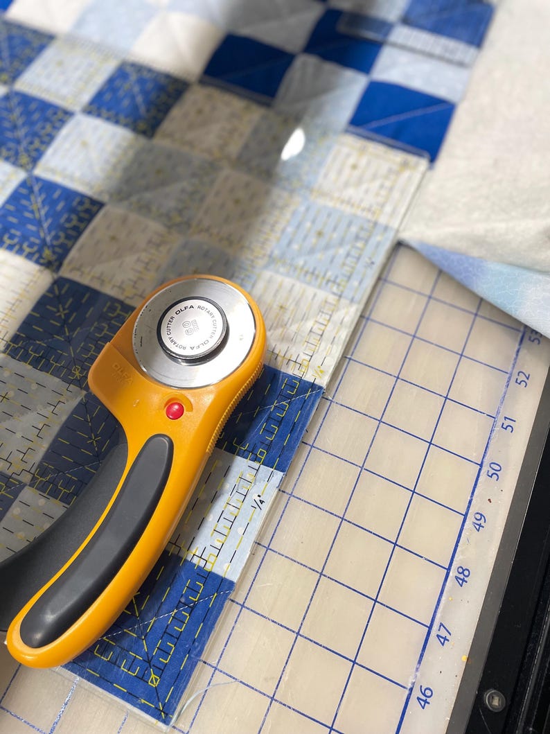 May include: An orange and black rotary cutter with a clear ruler is positioned on a blue and white checkered quilt. The ruler has measurements in centimetres. The quilt is in the process of being cut.