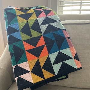 May include: A colorful quilt with a geometric pattern of triangles in black, pink, orange, yellow, green, and blue. The quilt is folded over a gray sofa, showcasing the intricate design and stitching.