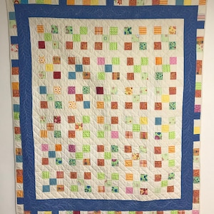 May include: A patchwork quilt with a blue border and a white background. The quilt is made up of small squares of different colours and patterns, including red, orange, yellow, green, blue, and pink.