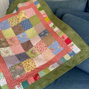 May include: A green patchwork quilt with a floral and geometric pattern. The quilt has a red border and a green binding. The quilt is folded and lying on a blue sofa.