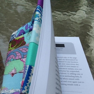 May include: A fabric book cover featuring a patchwork design of purple, teal, blue, and white floral and paisley patterns. The cover is wrapped around a white paper book with visible black text on the page, including the word sustainable.