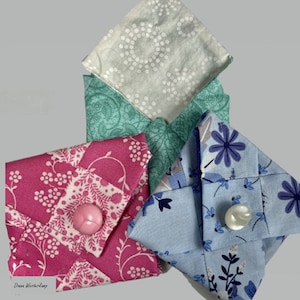 Può includere: Quattro quadrati di tessuto piegati in sacchetti con diversi motivi. I sacchetti sono realizzati in tessuto rosa, blu, verde e bianco con motivi floreali e geometrici. Un sacchetto ha un bottone rosa.