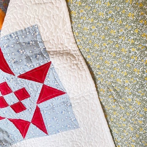 May include: A white quilt with a red and blue star pattern. The quilt is folded over a floral fabric with a green background.