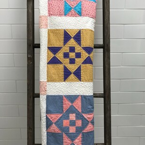 May include: A quilt with three different patterned squares. The top square is pink with blue and white stars. The middle square is yellow with purple stars. The bottom square is blue with pink stars.