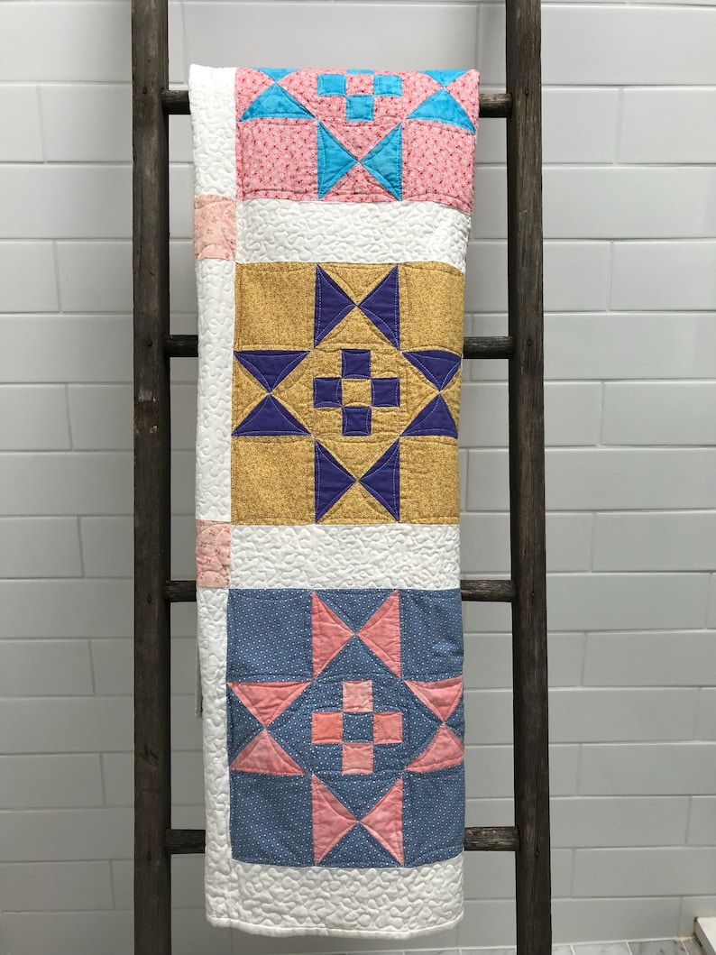 May include: A quilt with three different patterned squares. The top square is pink with blue and white stars. The middle square is yellow with purple stars. The bottom square is blue with pink stars.