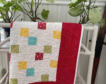 Könnte beinhalten: Eine weiße Steppdecke mit rotem Rand und einem Patchwork-Muster aus gelben, grünen und blauen Stoffquadraten. Die Steppdecke ist gefaltet und über einem weißen Holzgestell drapiert.