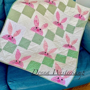 May include: A white and green checkered baby quilt with pink bunny faces. The quilt is folded on a blue sofa. The text "Donna Westerkamp" is visible in the bottom right corner.