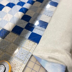 May include: A quilt in progress with a blue and white checkerboard design. A clear ruler and a rotary cutter are used for cutting the fabric. The quilt is on a white surface. The ruler shows measurements in inches.