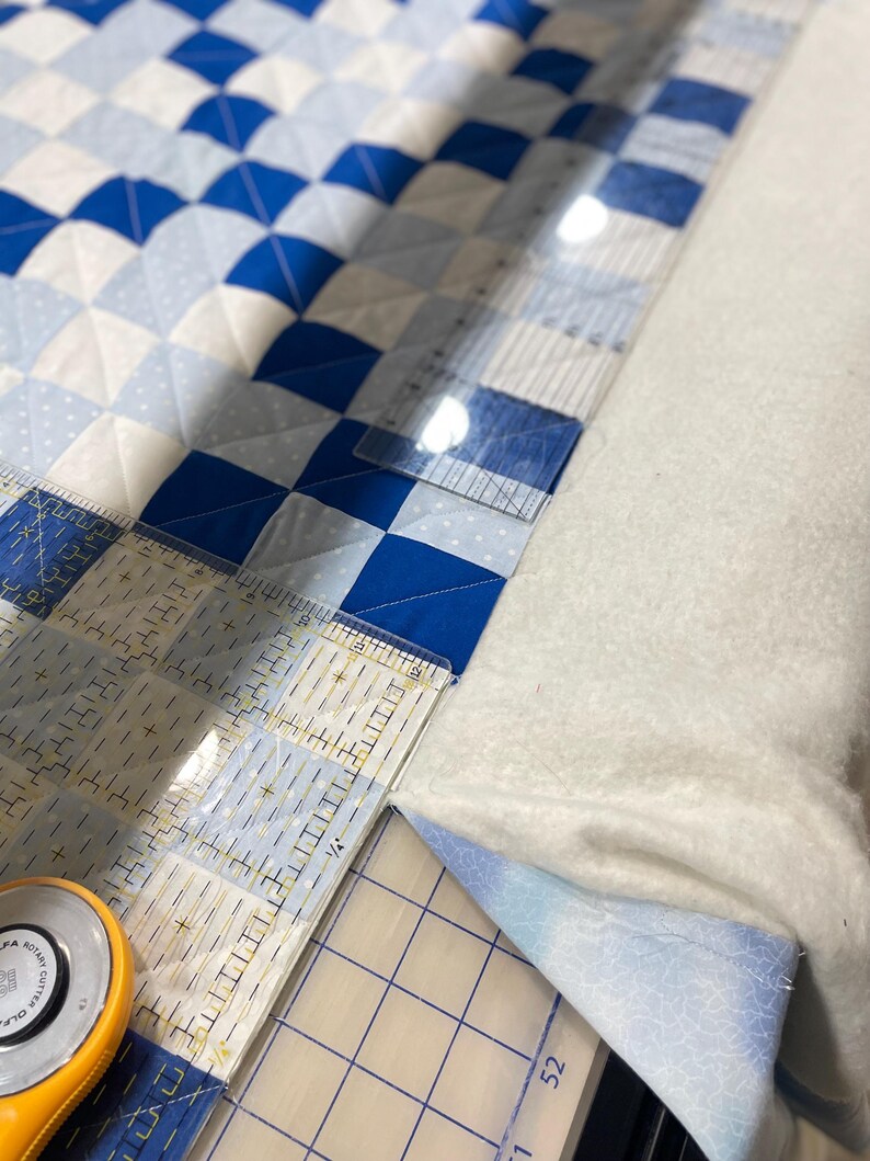 May include: A quilt in progress with a blue and white checkerboard design. A clear ruler and a rotary cutter are used for cutting the fabric. The quilt is on a white surface. The ruler shows measurements in inches.