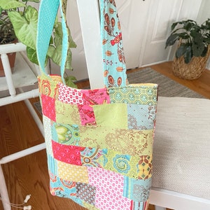 K&ouml;nnte beinhalten: Eine Patchwork-Tragetasche mit einem hellblauen und gr&uuml;nen Stoffgriff. Die Tasche besteht aus verschiedenen bunten Stoffen mit floralen und geometrischen Mustern.