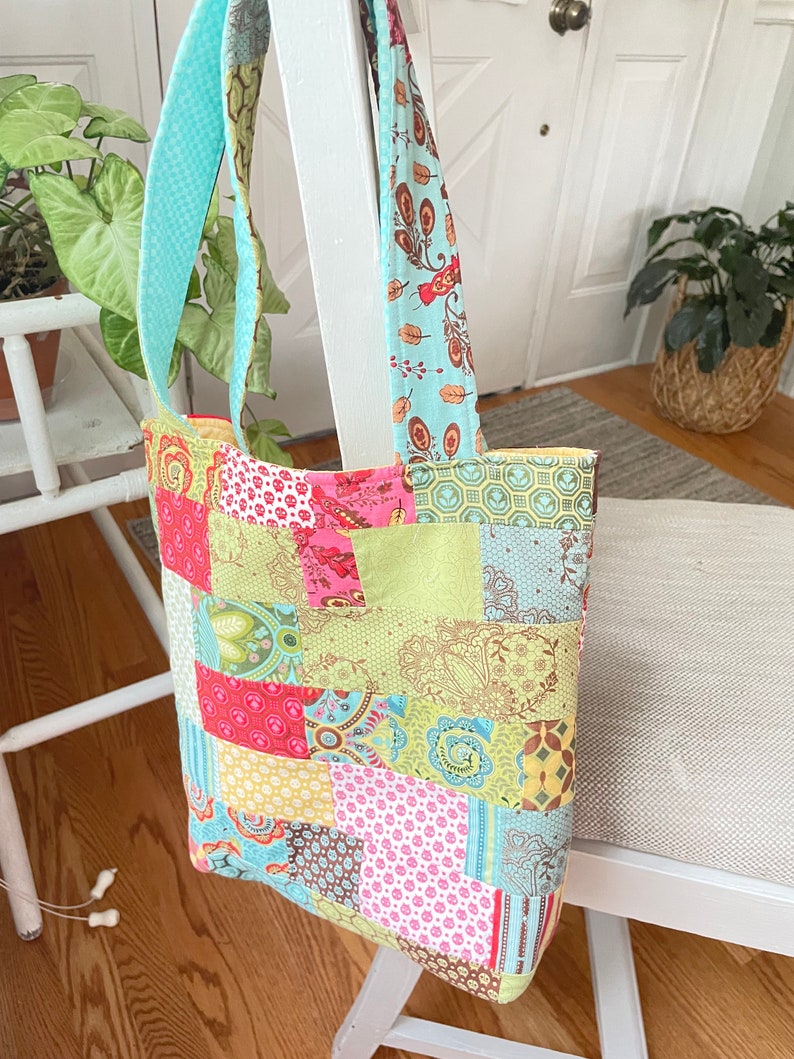 K&ouml;nnte beinhalten: Eine Patchwork-Tragetasche mit einem hellblauen und gr&uuml;nen Stoffgriff. Die Tasche besteht aus verschiedenen bunten Stoffen mit floralen und geometrischen Mustern.